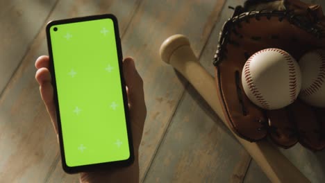 Person-Holding-Green-Screen-Mobile-Phone-Above-Baseball-Still-Life-With-Bat-Ball-And-Catchers-Mitt-On-Aged-Wooden-Floor-1